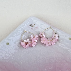 May include: A pair of gold hoop earrings adorned with delicate pink cherry blossom flowers. The earrings are set against a light pink and white background, showcasing the floral design. The earrings are a beautiful accessory.