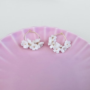 May include: A pair of gold hoop earrings adorned with clusters of white flower-shaped beads. The earrings are displayed on a pink, scalloped plate, creating a delicate and feminine aesthetic. The earrings are a floral accessory.