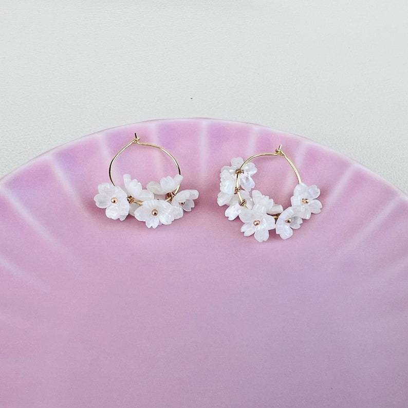 May include: A pair of gold hoop earrings adorned with clusters of white flower-shaped beads. The earrings are displayed on a pink, scalloped plate, creating a delicate and feminine aesthetic. The earrings are a floral accessory.