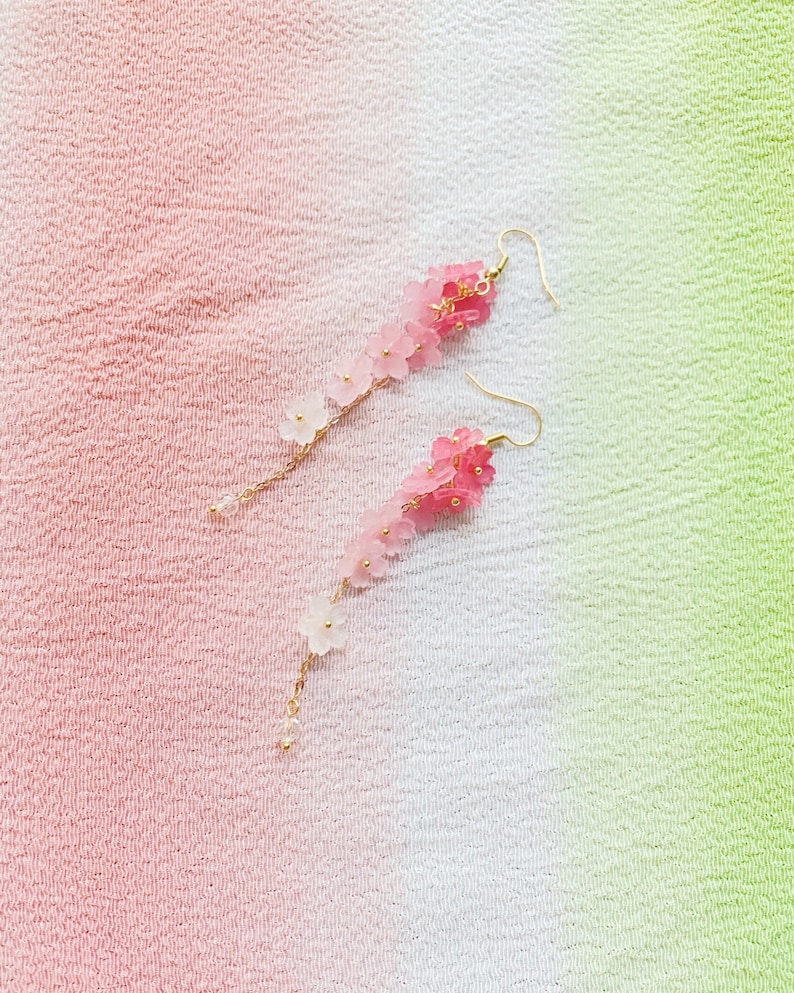 Puede incluir: Un par de aretes largos de flores rosadas y blancas. Los aretes est&aacute;n hechos de peque&ntilde;as flores de pl&aacute;stico enhebradas en una cadena dorada.