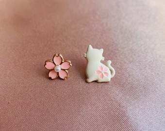 May include: A pair of earrings featuring a pink cherry blossom design with a pearl center and a white cat with a pink cherry blossom design.