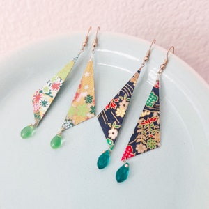 May include: Four origami-style earrings with floral patterns in green, yellow, and navy blue. Each earring has a teardrop-shaped green or teal bead. The earrings are displayed on a light blue plate.