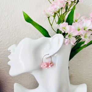 May include: A pair of gold hoop earrings adorned with clusters of small, light pink flowers. The earrings are displayed on a white mannequin head, with a backdrop of pink flowers and green stems.