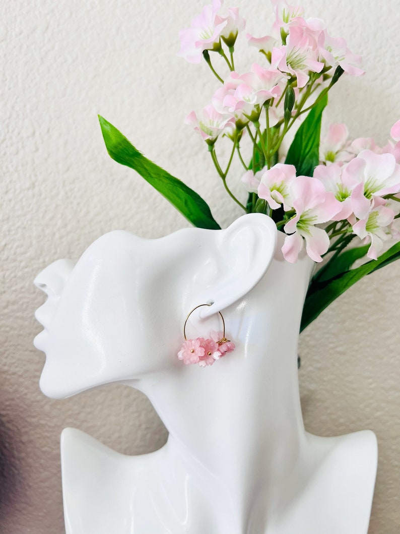 May include: A pair of gold hoop earrings adorned with clusters of small, light pink flowers. The earrings are displayed on a white mannequin head, with a backdrop of pink flowers and green stems.