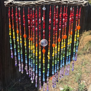 May include: A hanging decoration featuring a piece of textured, light-colored cholla wood at the top. Numerous strands of multi-colored beads in a rainbow gradient of red, orange, yellow, green, blue, and purple hang from the wood, with a large clear bead in the center.