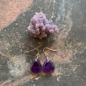 May include: A pair of teardrop-shaped amethyst earrings with silver hooks. The earrings are a deep purple color and are displayed with a cluster of light purple grape agate. The background is a textured, dark gray surface.
