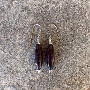 May include: A pair of earrings with elongated, oval-shaped, dark purple beads. The beads have a striped pattern and are suspended from silver-colored hooks. The earrings are displayed on a gray, textured surface.