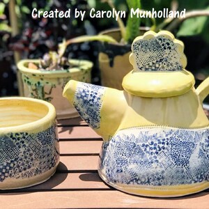 Może przedstawiać: Ż&oacute;łty czajnik i pasujący do niego dzbanek do śmietanki z niebieskimi i białymi wzorami. Czajnik ma ozdobną pokrywkę z falistym brzegiem.  "Stworzone przez Carolyn Munholland" jest napisane na obrazku.