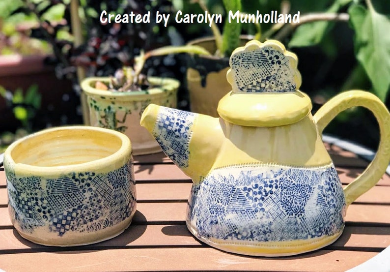 Może przedstawiać: Ż&oacute;łty czajnik i pasujący do niego dzbanek do śmietanki z niebieskimi i białymi wzorami. Czajnik ma ozdobną pokrywkę z falistym brzegiem.  "Stworzone przez Carolyn Munholland" jest napisane na obrazku.