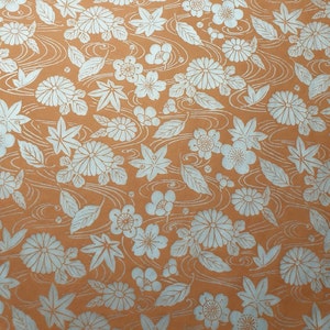 May include: A seamless pattern of white floral and leaf designs on a light orange background. The pattern features stylized flowers, leaves, and flowing lines.