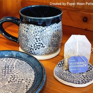 Może przedstawiać: Niebieski i biały kubek ceramiczny z dopasowanym spodkiem i małym spodkiem z torebką herbaty z napisem "Organic Blueberry Chamomile Herbal Tea".