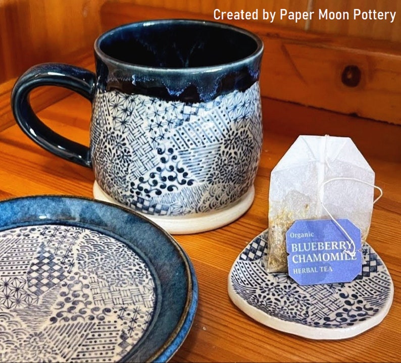 Może przedstawiać: Niebieski i biały kubek ceramiczny z dopasowanym spodkiem i małym spodkiem z torebką herbaty z napisem "Organic Blueberry Chamomile Herbal Tea".