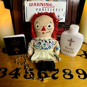 May include: A red-haired rag doll with a floral dress sits on a wooden surface with a small book, a rosary, and a bottle of holy water. The doll has a warning sign that reads "POSITIVELY DO NOT OPEN".