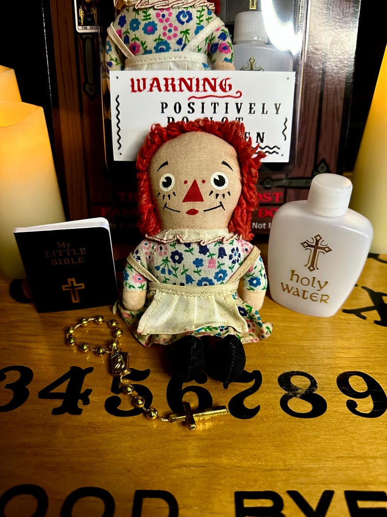 May include: A red-haired rag doll with a floral dress sits on a wooden surface with a small book, a rosary, and a bottle of holy water. The doll has a warning sign that reads "POSITIVELY DO NOT OPEN".