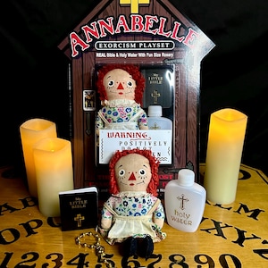 May include: A vintage-style doll with red hair and a floral dress sits on a wooden Ouija board with a small bible, a bottle of holy water, and a rosary. The doll is part of an Annabelle Exorcism Playset.