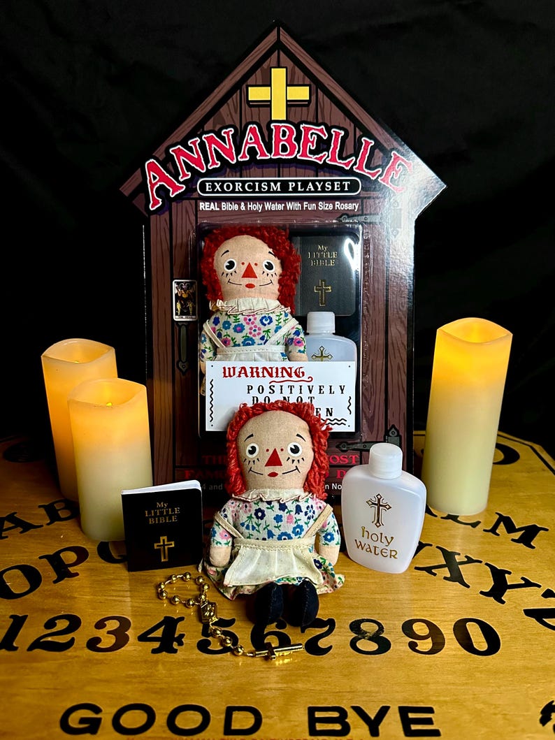May include: A vintage-style doll with red hair and a floral dress sits on a wooden Ouija board with a small bible, a bottle of holy water, and a rosary. The doll is part of an Annabelle Exorcism Playset.
