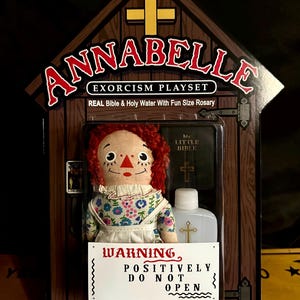 May include: A red-haired doll with a floral dress and a white apron is displayed in a wooden box with a cross on the front. The box also contains a small bible and a bottle of holy water. The text on the box reads "Annabelle Exorcism Playset" and "Warning Positively Do Not Open".