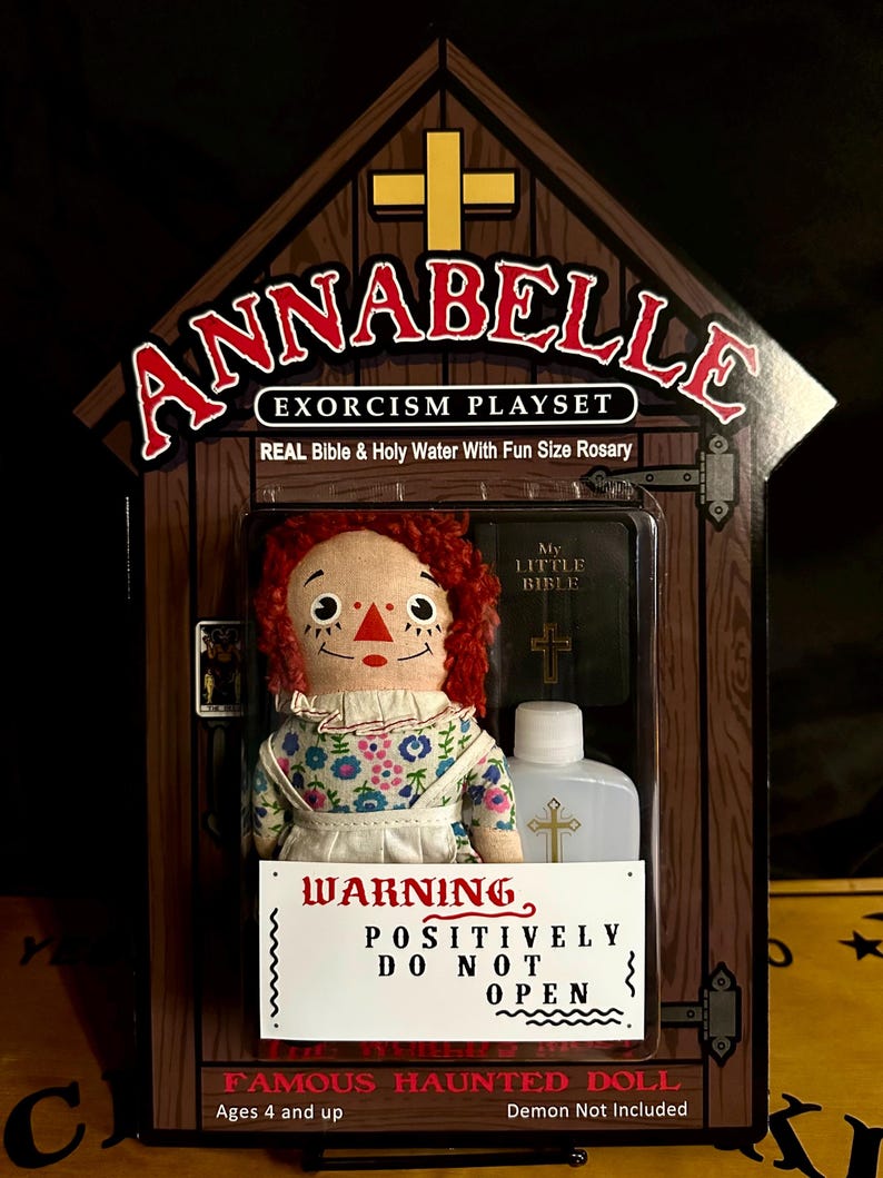May include: A red-haired doll with a floral dress and a white apron is displayed in a wooden box with a cross on the front. The box also contains a small bible and a bottle of holy water. The text on the box reads "Annabelle Exorcism Playset" and "Warning Positively Do Not Open".