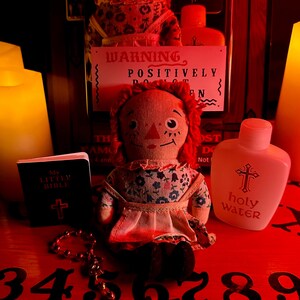 May include: A red Ouija board with black numbers 3 through 9, a small red and white doll, a small book with a cross on the cover that says "My Little Bible", a bottle of holy water with a cross on the label, and a string of beads with a cross charm.
