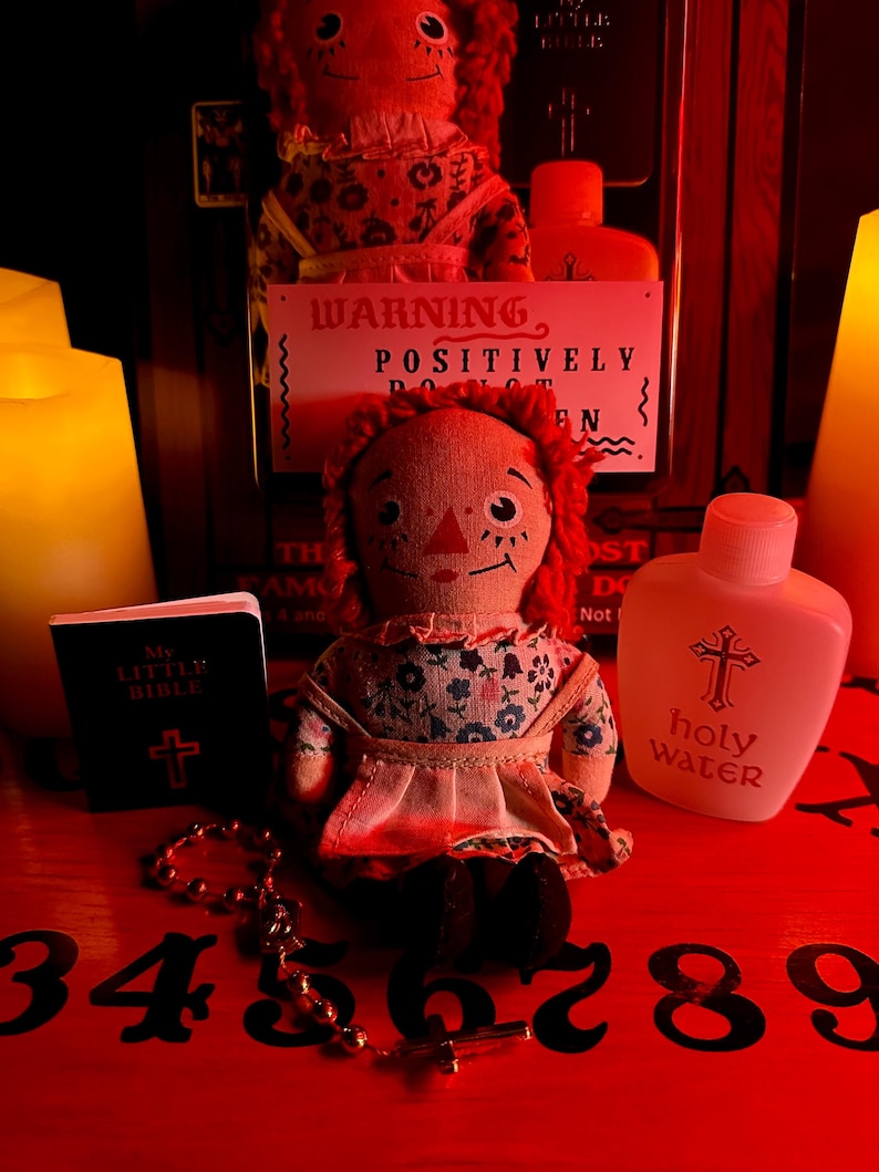 May include: A red Ouija board with black numbers 3 through 9, a small red and white doll, a small book with a cross on the cover that says "My Little Bible", a bottle of holy water with a cross on the label, and a string of beads with a cross charm.