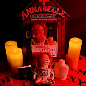 May include: A red and white cardboard box with the words "Annabelle Exorcism Playset" on the front. The box contains a doll, a small bottle of holy water, a small bible, and a rosary. The box is sitting on a red and black Ouija board.