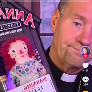 May include: A man wearing a black clergy shirt with a white collar holds a red-haired doll in a glass case. The doll is wearing a white dress with floral patterns. The case has a label that reads "REAL Bible & Holy Water" and "EXORCISM". The man is looking at the camera with a serious expression. The text "THERE'S NO WAY. THIS CAN'T BE REAL. IT MUST BE A JOKE... IT'S NOT." is displayed above the image. The text "BISHOP JAMES LONG, D. MIN, OSB" is displayed below the image.