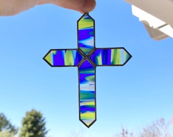 May include: A stained glass cross with blue, green, and yellow swirls. The cross is hanging from a window.
