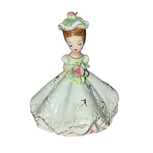 May include: A vintage porcelain figurine of a woman in a long, flowing dress. The figurine has a light blue dress with gold trim and a matching hat. She is holding a bouquet of pink roses. The figurine is approximately 15 cm tall.