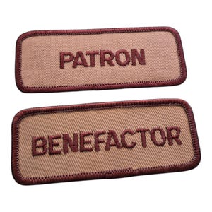 May include: Two rectangular patches with embroidered text. The top patch reads "PATRON" in maroon thread on a tan background, framed by a maroon border. The bottom patch reads "BENEFACTOR" in the same style.
