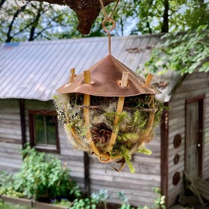 May include: A copper birdhouse with a woven nest made of twigs, moss, and pine cones. The birdhouse is hanging from a tree branch.