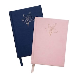 May include: Two hardcover journals, one navy blue and one pink, are displayed. Each journal features a gold leaf design in the upper right corner and a matching ribbon bookmark. The journals are set against a white background.