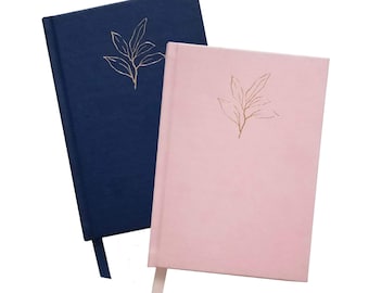 May include: Two hardcover journals, one navy blue and one pink, are displayed. Each journal features a gold leaf design in the upper right corner and a matching ribbon bookmark. The journals are set against a white background.