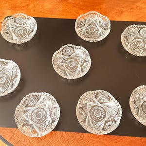 Könnte beinhalten: Neun klare, geschliffene Glasschalen mit Stern- und Blumenmustern sind auf einer schwarzen Oberfläche angeordnet. Die Schalen haben gewellte Ränder und dienen wahrscheinlich zum Servieren von Desserts oder kleinen Portionen.