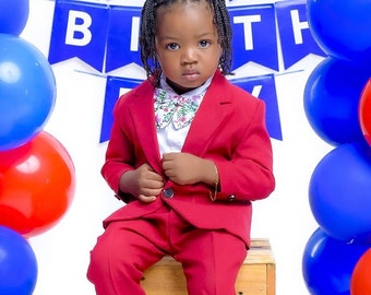 May include: A young child wearing a red suit and white sneakers sits on a wooden crate in front of a blue and white "Happy Birthday" banner. Blue and red balloons are on either side of the child.