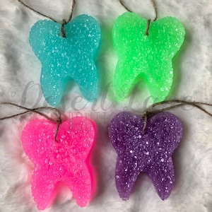以下が含まれることがあります： 青、緑、ピンク、紫の4つの歯の形をした芳香剤。各歯はキラキラした質感のある素材でできており、茶色の紐で吊り下げられています。