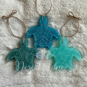 May include: Three teal and blue turtle-shaped air fresheners with a sparkly finish. Each turtle is attached to a twine string with a bow.