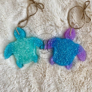 May include: Two teal and purple turtle-shaped car air fresheners with glitter embedded in the resin. The turtles are hanging from twine.