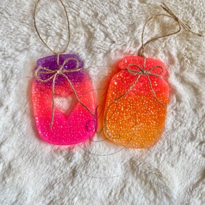 May include: Two colorful glass jar-shaped ornaments with a heart cut-out. The jars are filled with colorful beads and have a rustic twine bow.