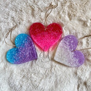 May include: Three heart-shaped car air fresheners in pink, purple, and white with a glittery finish. The hearts are hanging from a string.