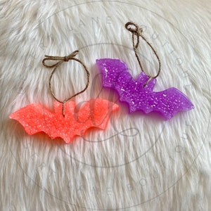 May include: Two hanging bat-shaped air fresheners. One is a bright orange color and the other is a bright purple color. Both are made of a glittery material.
