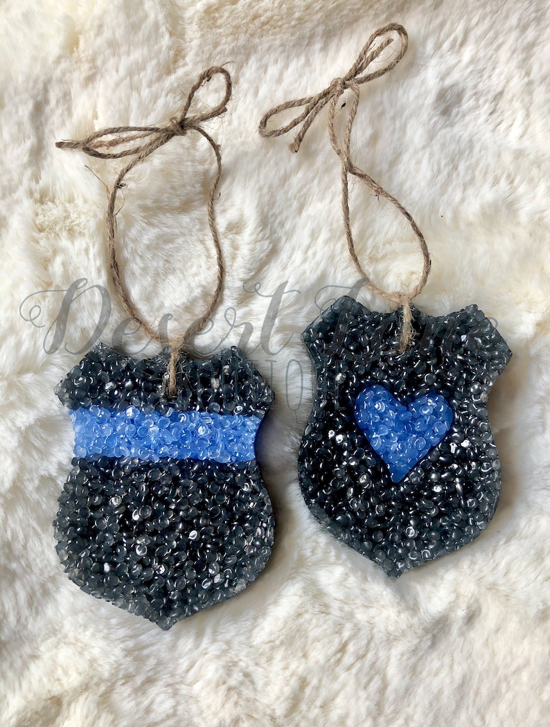 May include: Two black and blue police badge-shaped car air fresheners with a blue ribbon and a blue heart. The air fresheners are hanging from twine.