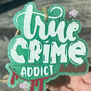 May include: Green and white true crime addict car air freshener with a red knife and dripping blood. The air freshener is hanging from a brown twine string.