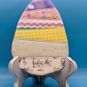 Puede incluir: Decoración de huevo de Pascua de tela colorida. El huevo está hecho de varias telas estampadas, incluyendo cuadros, flores y lunares. Se encuentra sobre un soporte blanco decorativo con un estampado floral y de conejitos.