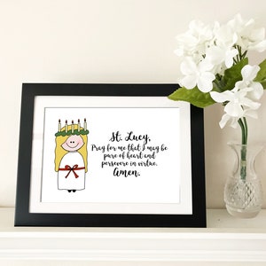 May include: A framed print with a black frame and a white background. The print features a cartoon illustration of a woman wearing a white dress and a wreath of candles on her head. The text below the illustration reads "St. Lucy, Pray for me that I may be pure of heart and persevere in virtue. Amen."