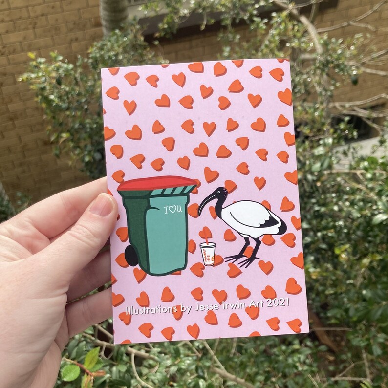 May include: A pink card featuring a white ibis bird next to a green bin with the words "I love you" written on it. The background is covered in small red hearts. The card reads "Illustrations by Jesse Irwin Art 2021".