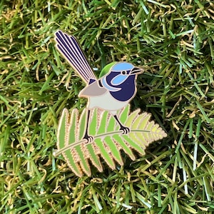 May include: An enamel pin featuring a blue and white bird perched on a green fern leaf. The bird has a long tail and a blue head with a white stripe.