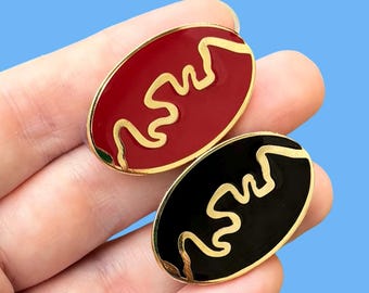 May include: Two enamel pins with gold outlines. One pin is red with a gold wavy line design. The other pin is black with a gold wavy line design.