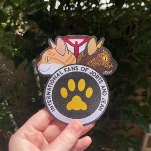 May include: A black and white embroidered patch featuring a paw print in the centre. The text surrounding the paw print reads "International Fans of Jorts and Jean". The patch is bordered by two cartoon cats, one orange and one brown.