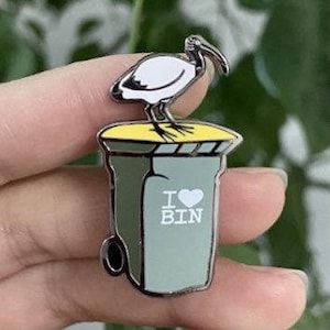 May include: An enamel pin depicting a white ibis bird standing on a green bin with the words "I ♥ BIN" on the side.