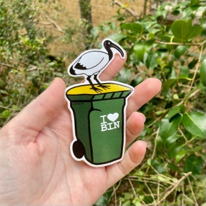 May include: A green and yellow bin sticker with a white ibis bird standing on top. The bin has the words "I love bin" with a red heart.